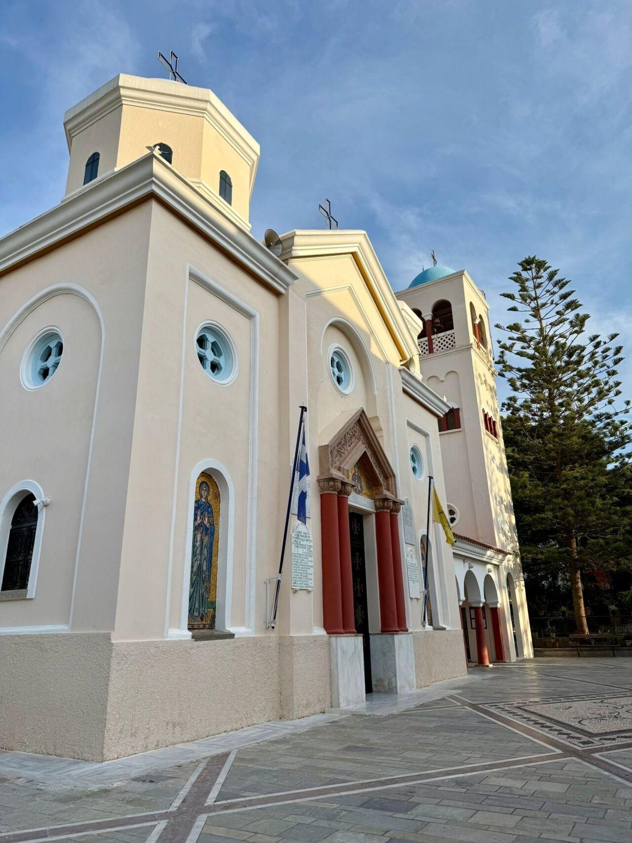 Eine hellgelbe Kirche mit Rundfenstern, roten Türen und Glockenturm in klarem Abendlicht.