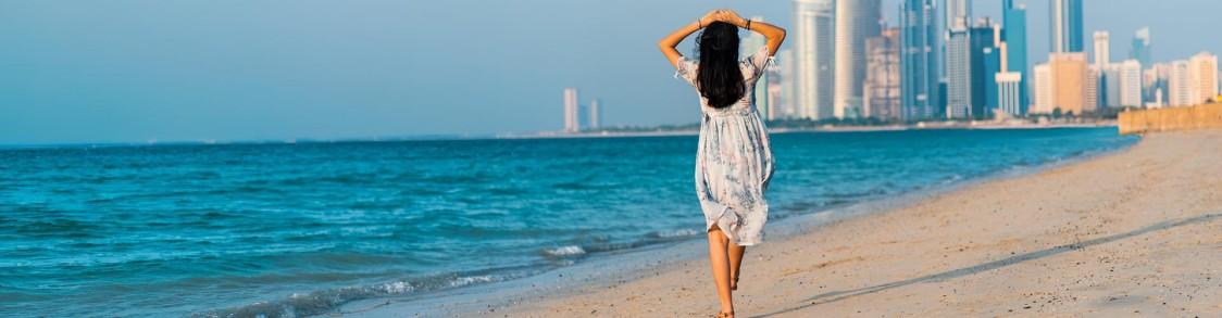 Frau im hellen Kleid geht am Sandstrand, Blick auf moderne Skyline und ruhiges Meer bei Tageslicht.