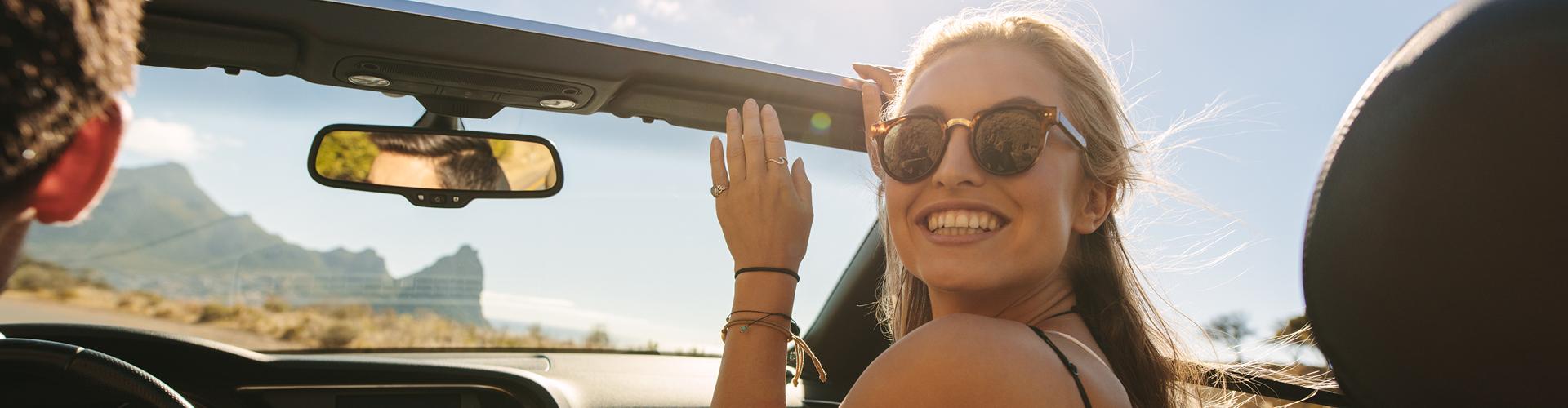 Junge Frau mit Sonnenbrille fährt Cabrio, sonnige Landschaft mit Bergen im Hintergrund, Urlaubsstimmung.