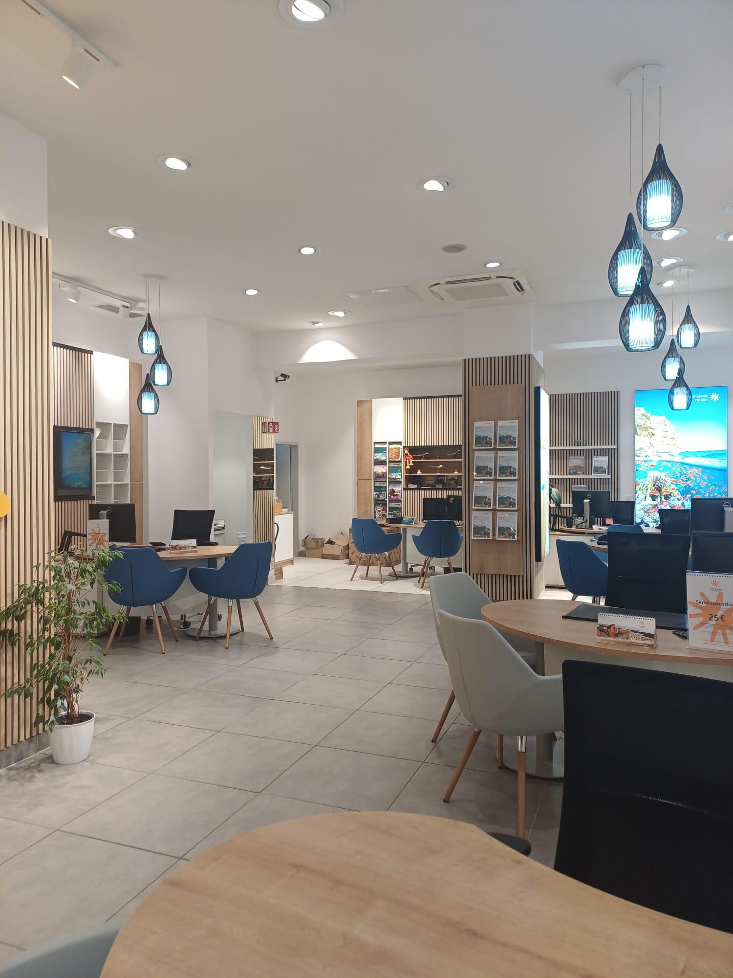 Helles, modernes Reisebüro mit blauen Sesseln, hängenden blauen Lampen und naturfarbenen Holztischen.
