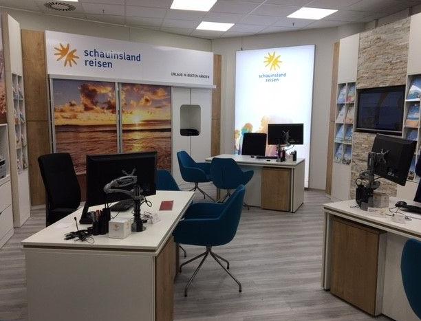 Modernes Reisebüro mit hellen Holzmöbeln, blauen Stühlen und Urlaubsbildern an den Wänden.