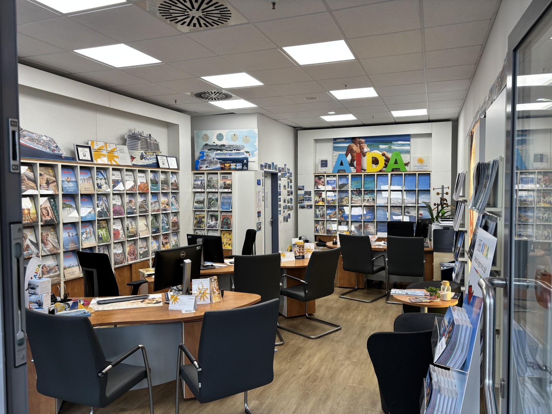 Moderner Reisebüro-Showroom mit Regalen voller Reisekataloge, mehreren Tischen und schwarzen Stühlen.