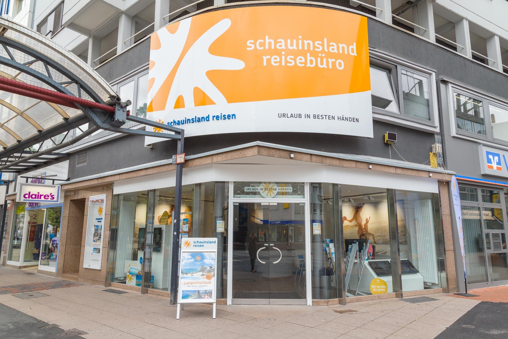 Reisebüro mit großem orangeweißem Schild, Glasfenstern, modernen Eingängen und nahegelegenen Geschäften.