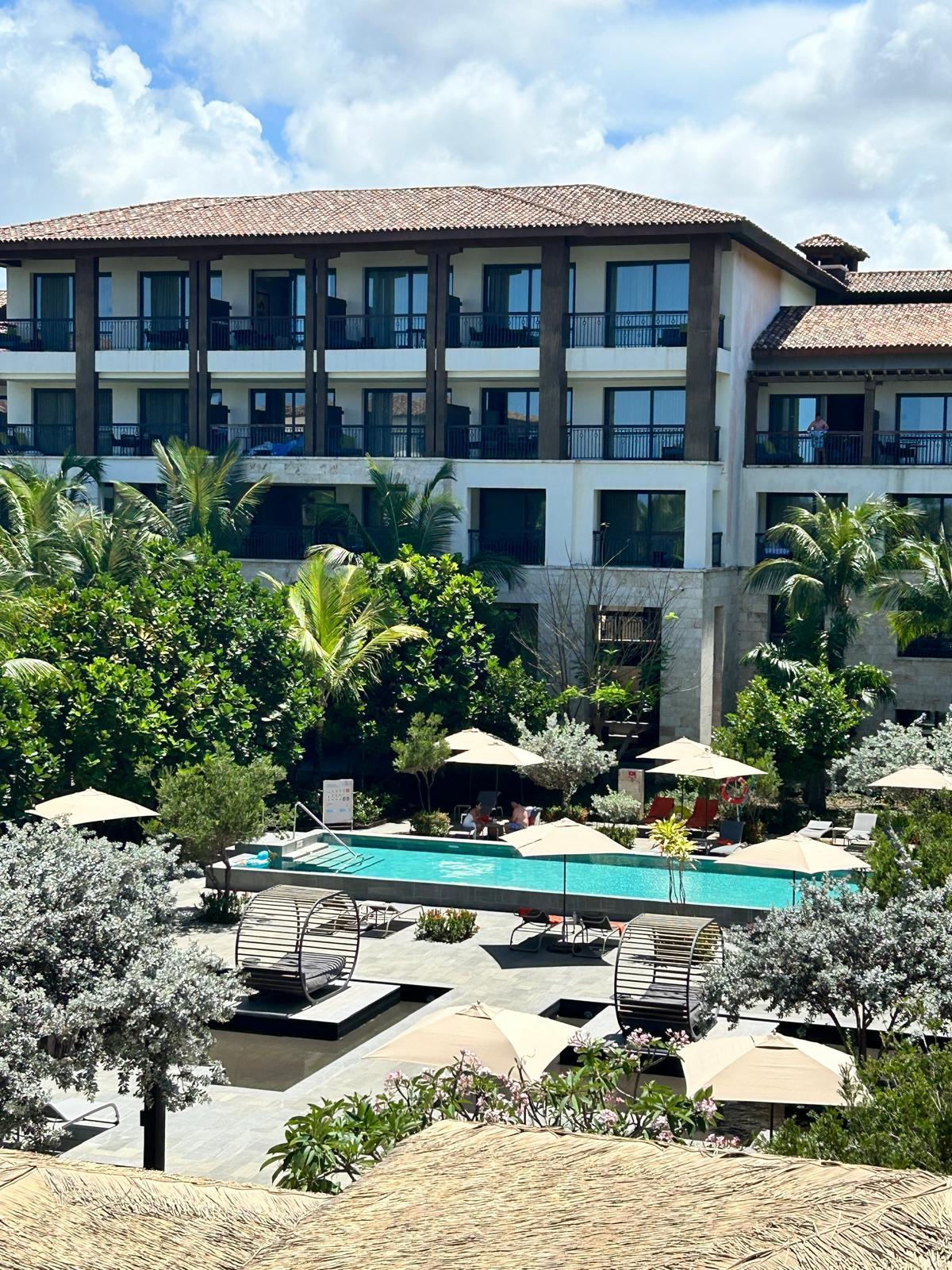 Luxuriöses Hotel mit Pool, Liegestühlen und grüner tropischer Gartenanlage unter blauem Himmel.