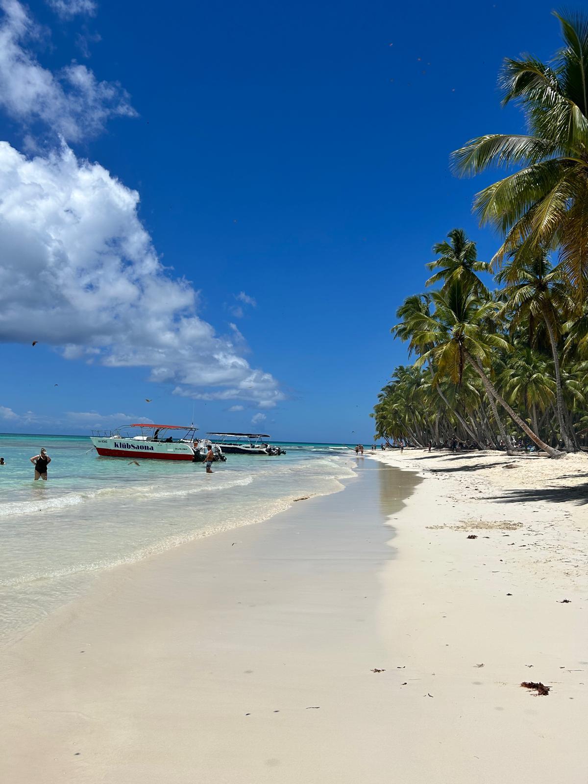 Weißer Sandstrand mit Palmen, klarem türkisfarbenem Wasser und Booten unter blauem Himmel.