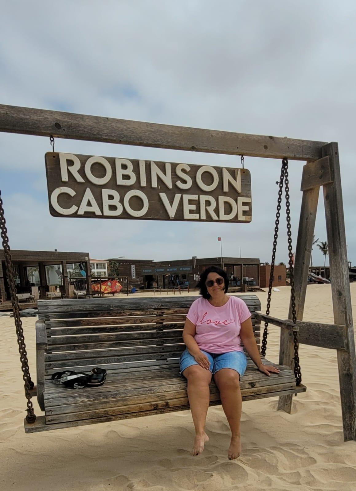 Frau entspannt auf großer Holzschaukel im Sand vor dem Robinson Cabo Verde Hotel bei bewölktem Himmel.