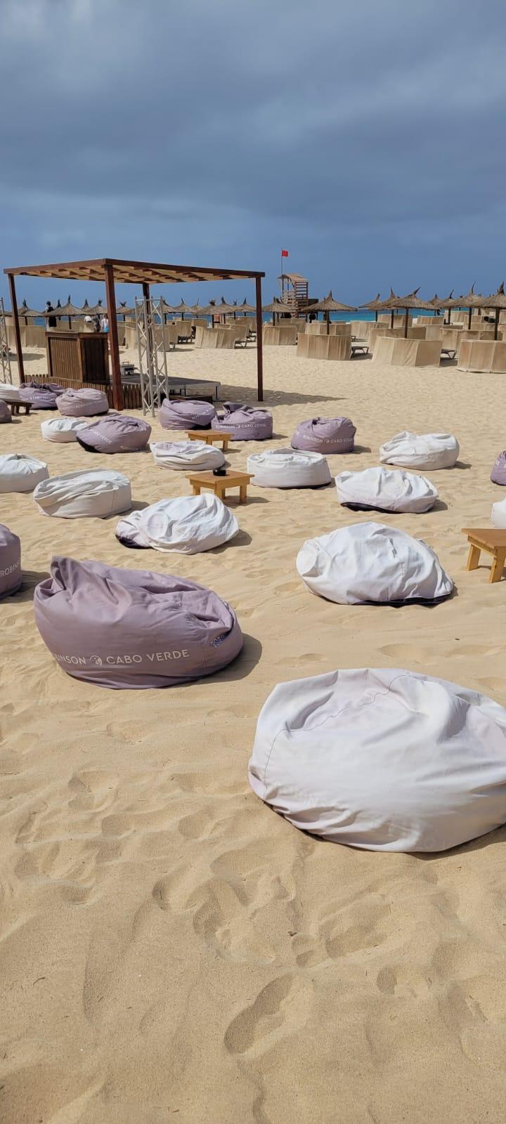 Sandstrand mit weißen und lilafarbenen Sitzsäcken, Holzhütten und Sonnenschirmen unter bewölktem Himmel.