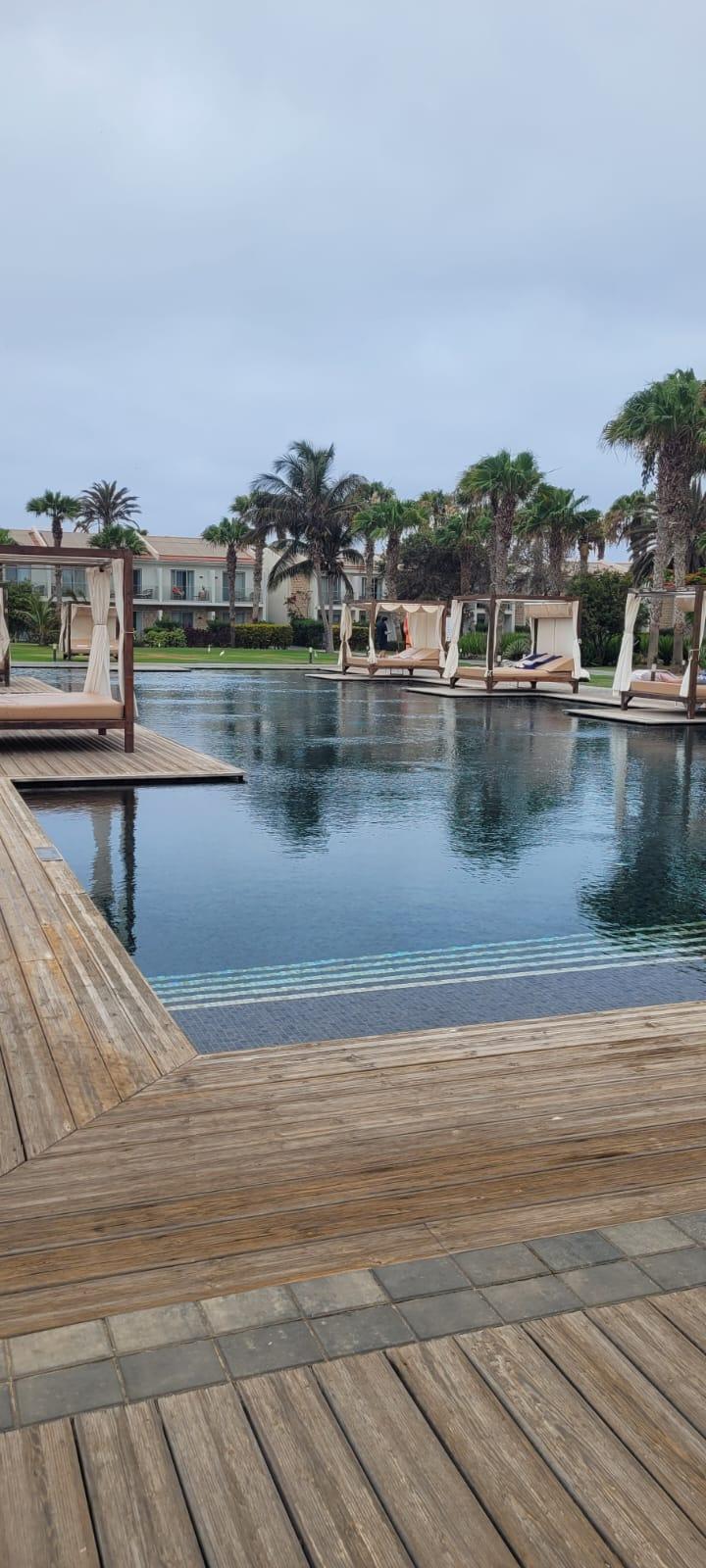 Moderner Hotelpool mit ruhigem Wasser, Holzdeck und überdachten Liegen, umgeben von Palmen.